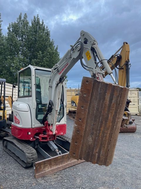 Mini excavator lifting a rusty metal panel in a gravel yard, with construction equipment in the background