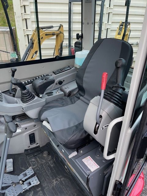 Compact excavator cab with gray seat, control levers, and red safety lever inside the operator area