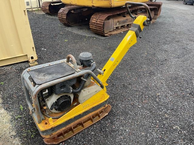 Yellow walk-behind construction compactor on gravel, with tracked machinery in the background