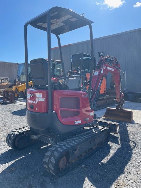 Red mini excavator on gravel in a sunny industrial yard.