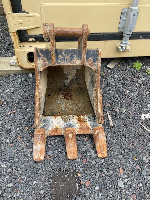 Rusty excavator bucket with teeth resting on gravel beside a yellow machine.