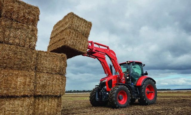 A red tractor is loading bales of hay into a pile.