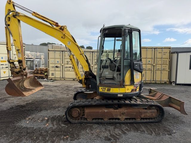 Yellow excavator on tracks in a construction yard, with shipping containers in the background.