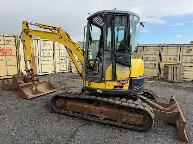 Yellow excavator on tracks in a storage yard, with shipping containers and buckets nearby.