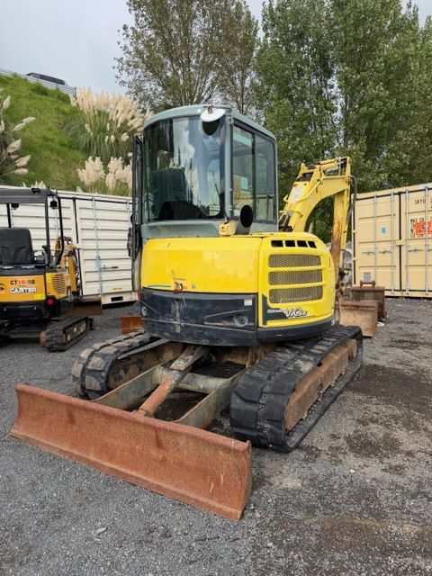 Yellow tracked excavator with front blade parked on gravel in a yard, with trucks and trees behind it
