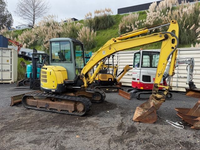 Yellow mini excavator on tracks parked in a yard beside shipping containers and metal debris