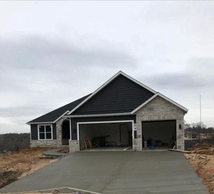 Newly built house with gray siding, stone facade, and a wide concrete driveway under an overcast sky