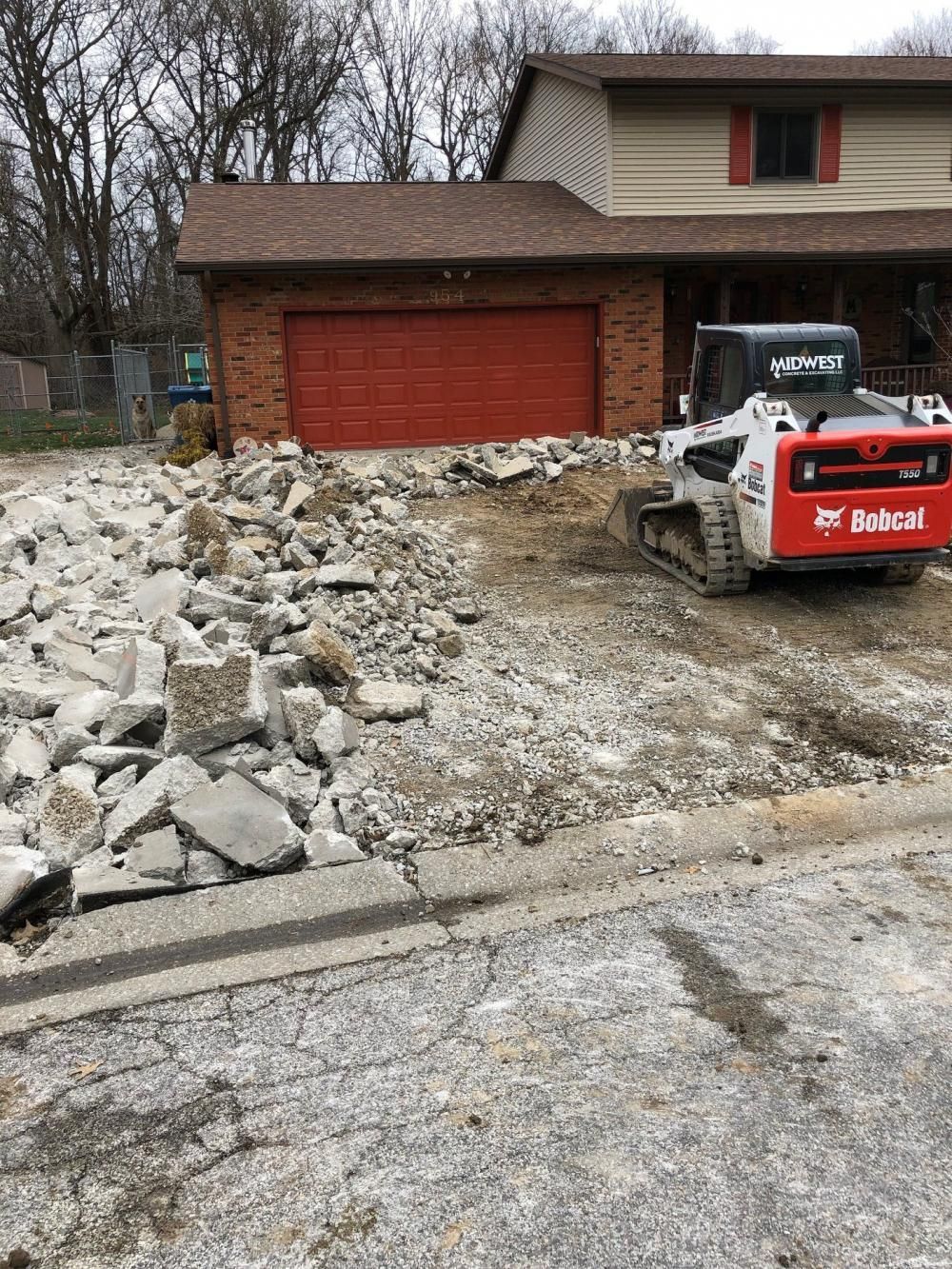 Bobcat excavator beside rubble on a driveway in front of a brick house with a red garage door.