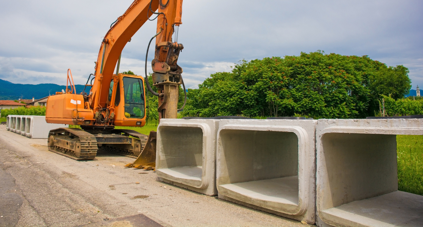 Excavator lifting a concrete drainage section beside stacked culvert blocks on a roadside construction site