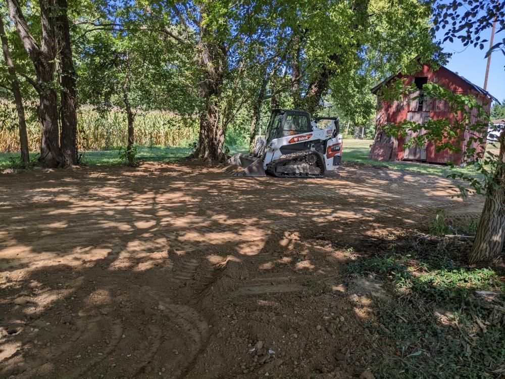 White SUV parked on a dirt clearing beside a red barn, with trees casting shade around it.