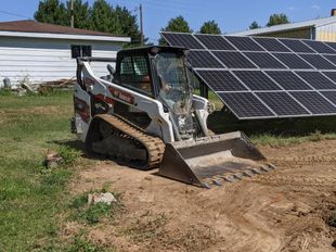 White skid steer with bucket on dirt beside solar panels near a house and trees