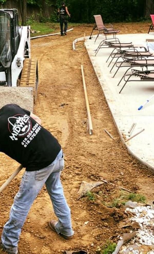 Worker leveling dirt beside a pool patio with lounge chairs on the right.