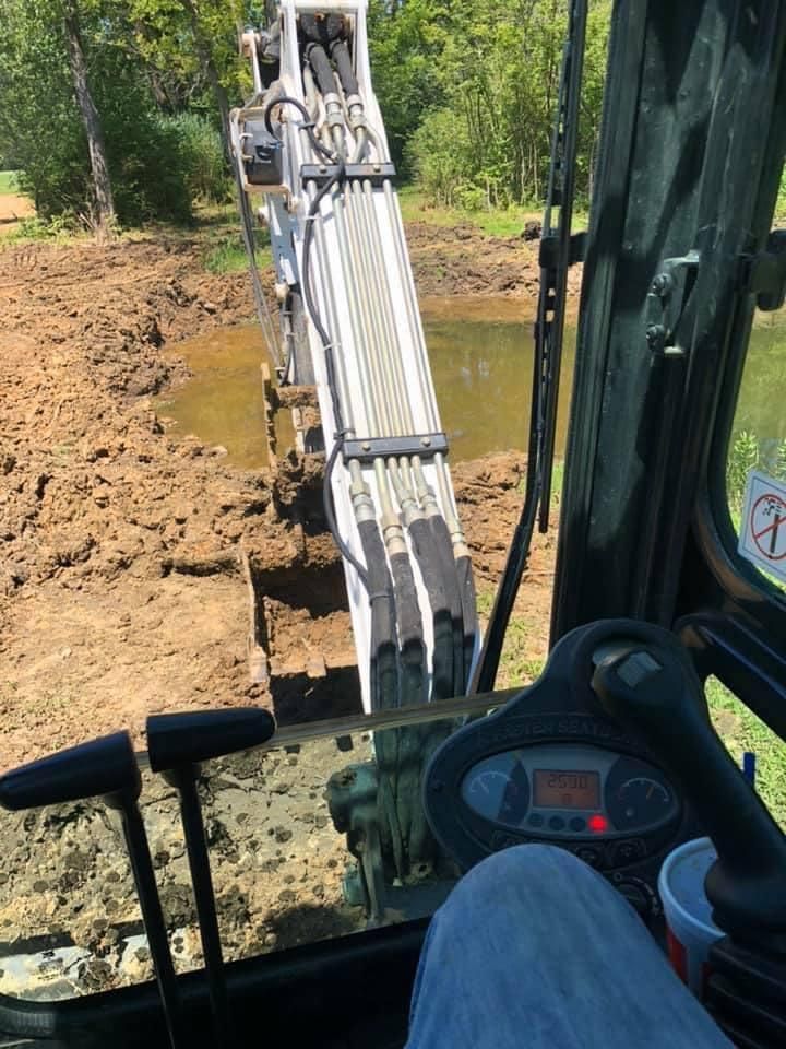Inside an excavator cab, the arm digs a muddy trench beside a wooded area