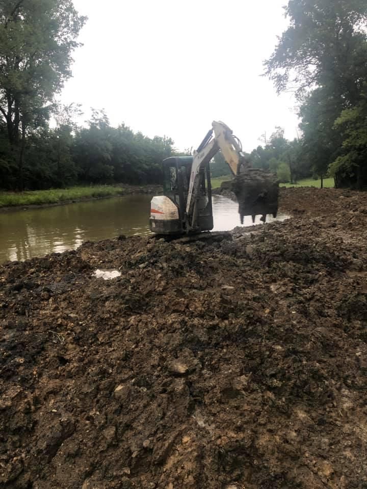 Excavator digging mud beside a river under cloudy skies, surrounded by trees