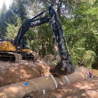 Excavator lifting a large concrete pipe in a wooded construction area