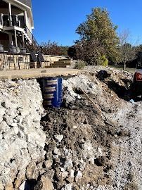 Construction trench beside house with exposed dirt, a blue barrel, and a tree under a clear sky