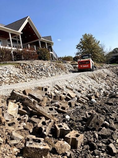 Small red excavator on a rocky construction site beside a house under a clear blue sky