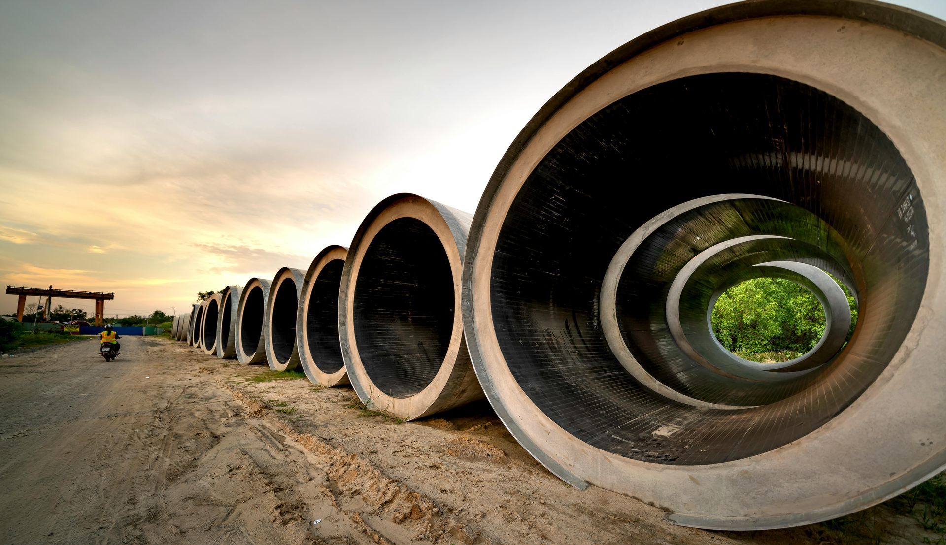 Row of large concrete pipes receding into the distance along a sandy construction site at sunset