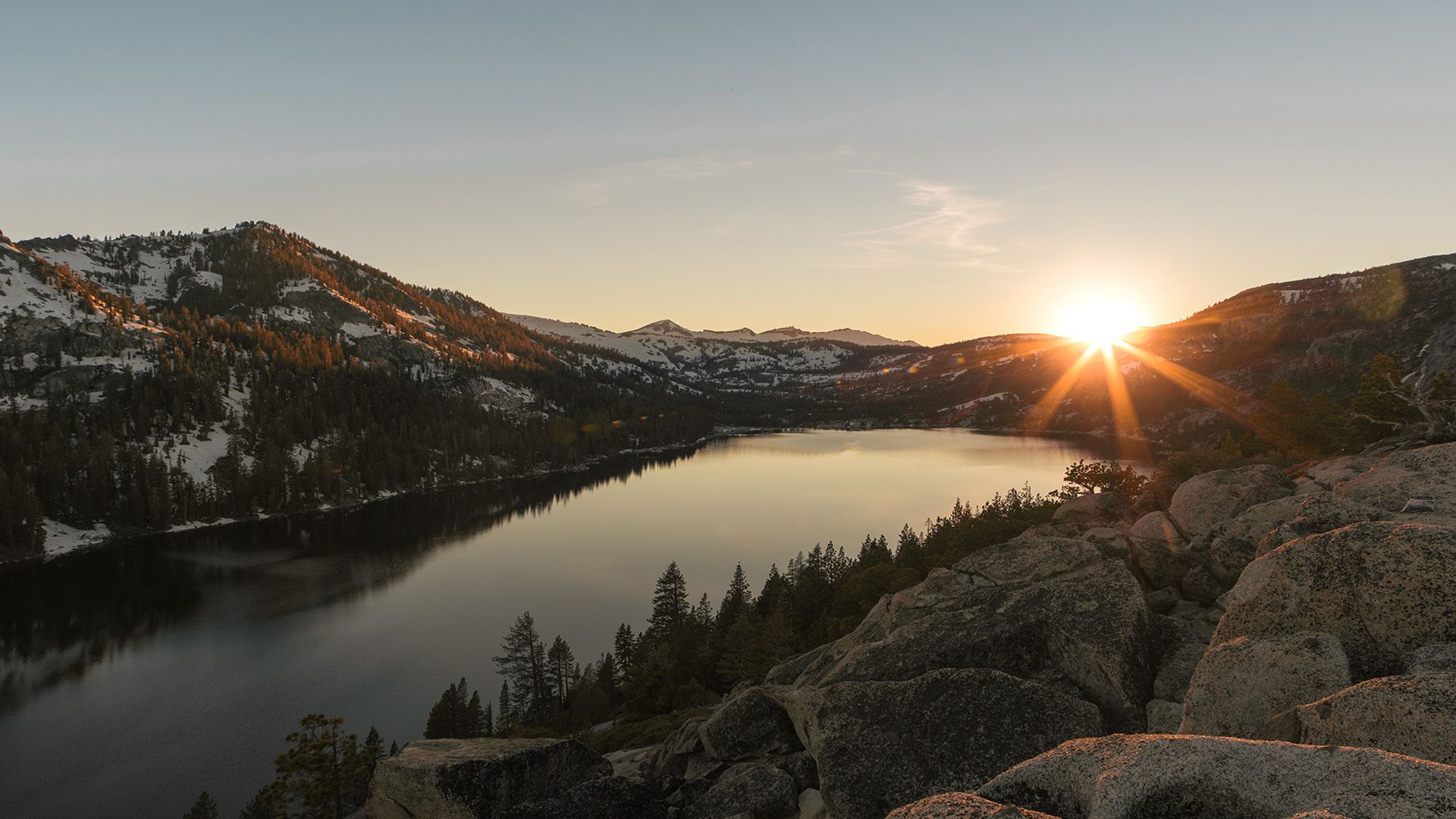 Sunset over a mountain lake, with snow patches, trees, and rocky foreground.