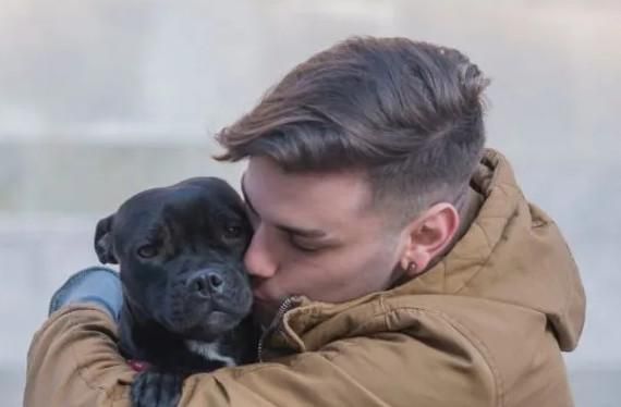 Man kisses a black dog, both are outdoors, man in brown jacket.