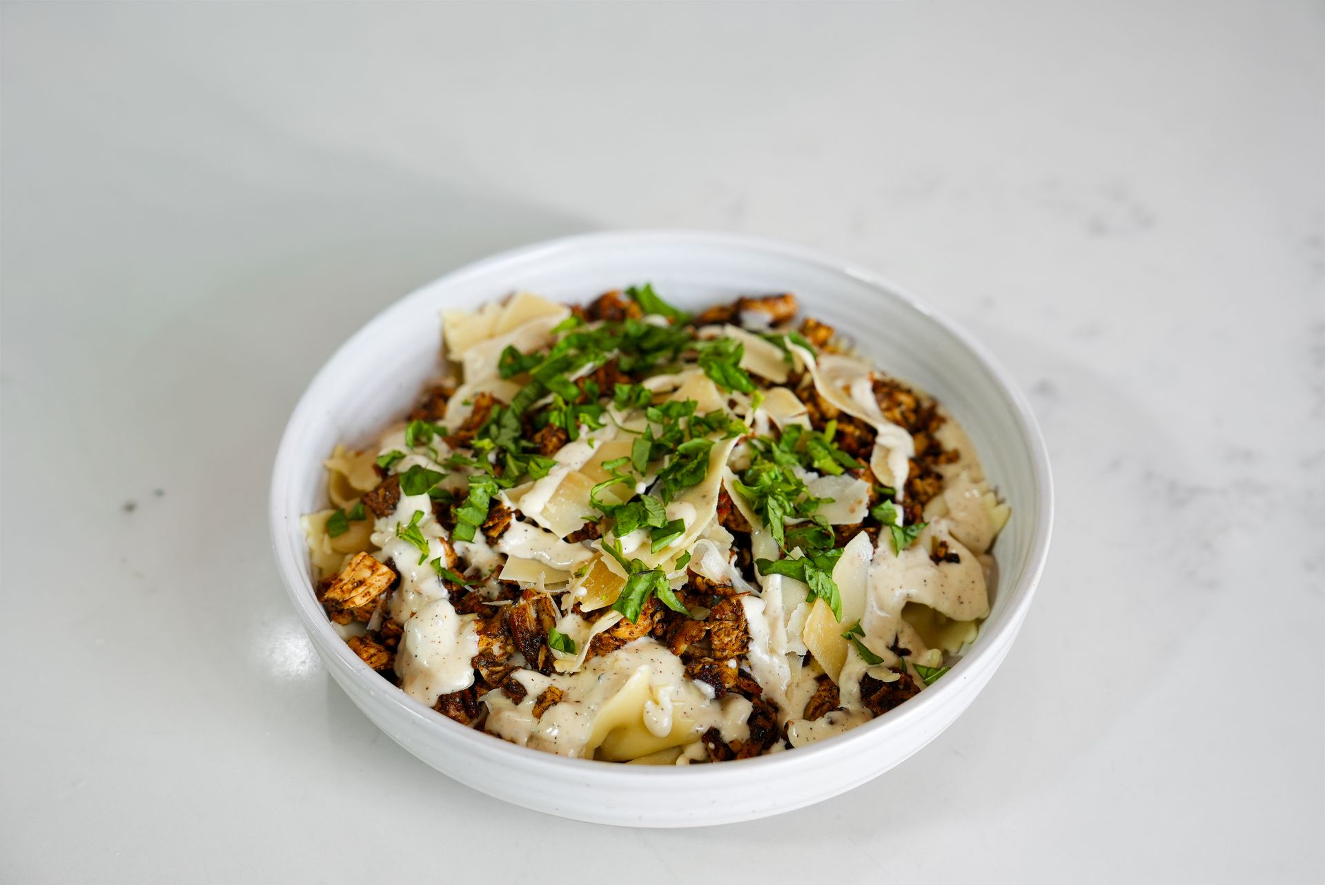 Bowl of pasta topped with shaved cheese, herbs, and a dark sauce on a white table.