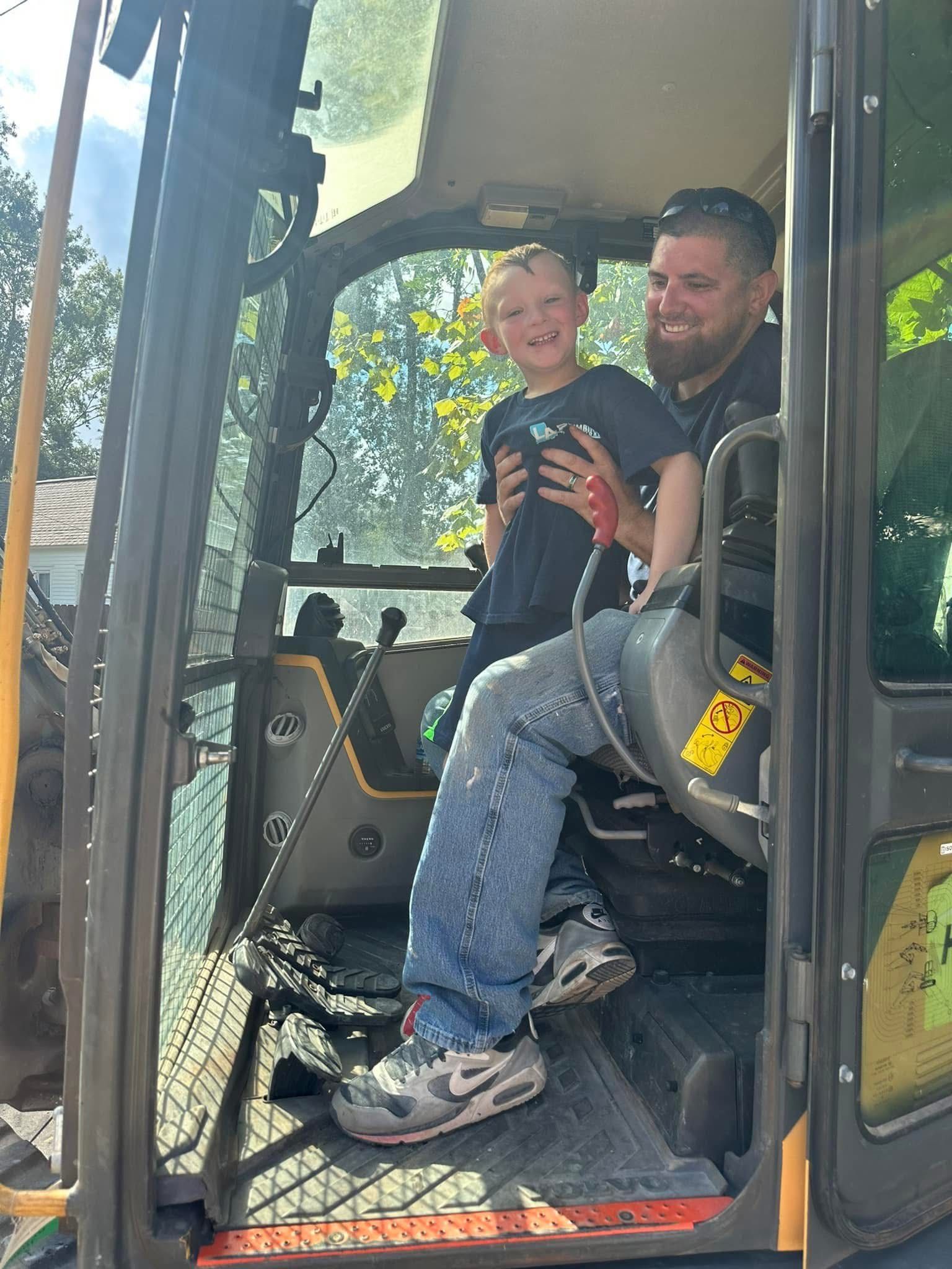 Man and young boy in a construction vehicle cab; boy smiles, man holds him; sunny outdoor setting.