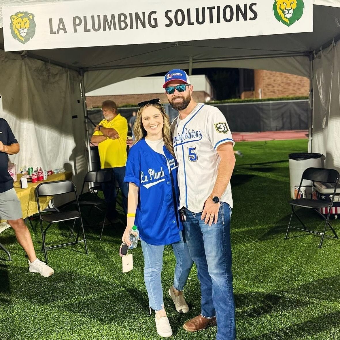 Two people smiling, wearing baseball jerseys, posing in front of a tent with