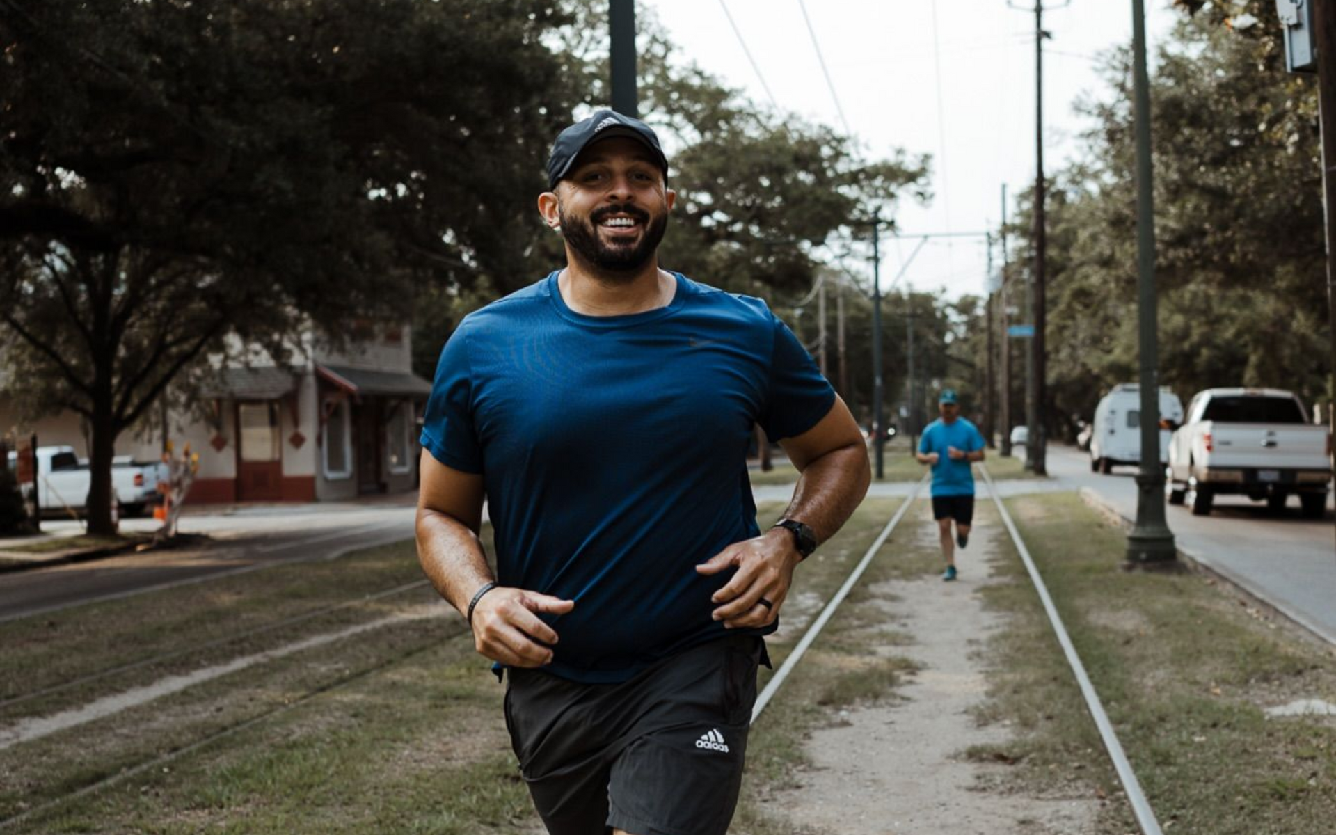 A smiling runner in a blue shirt and dark shorts jogging on a path with a second person running in the distance.