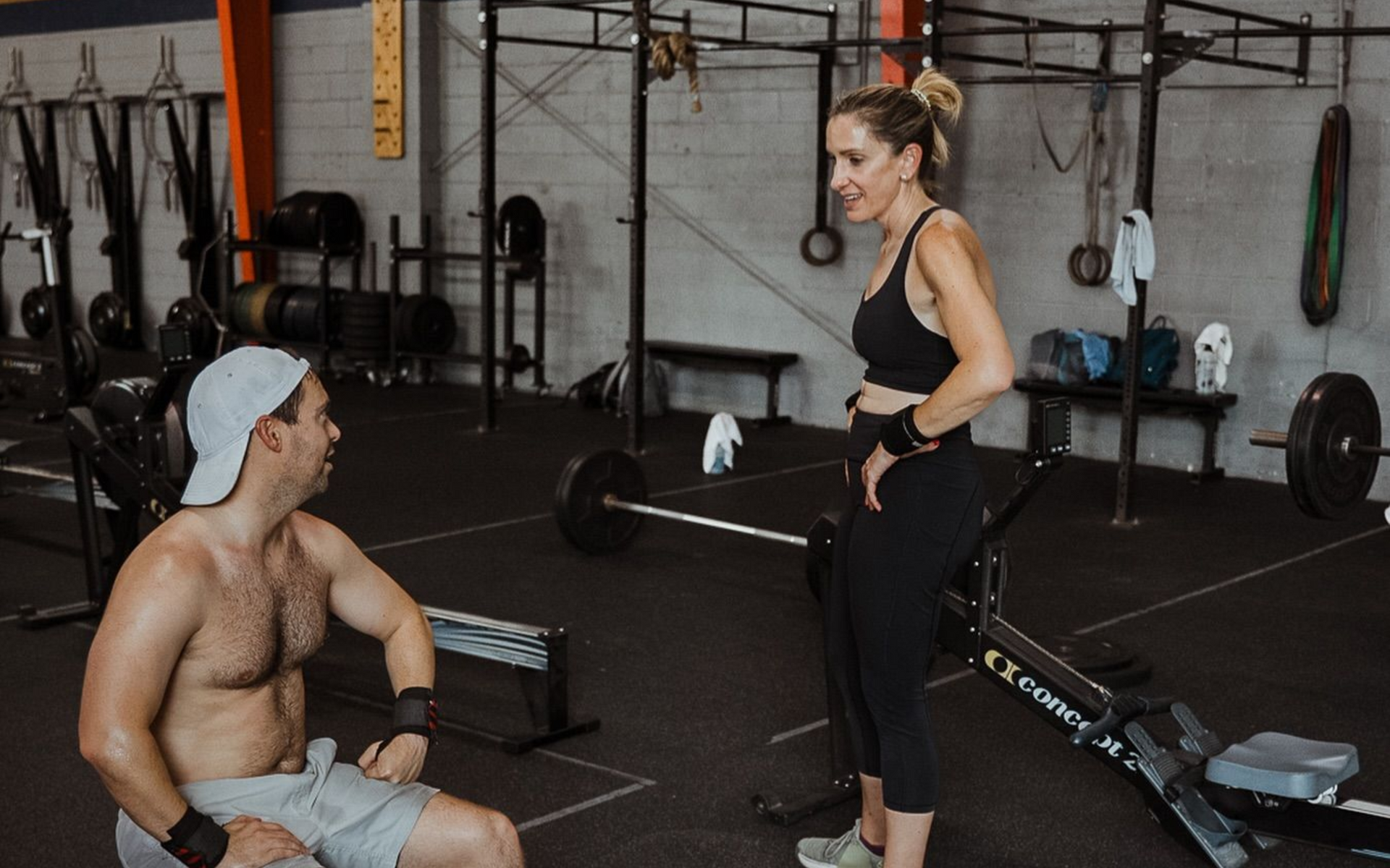 A person in a gym in New Orleans sits on a bench talking to another person standing next to a rowing machine.