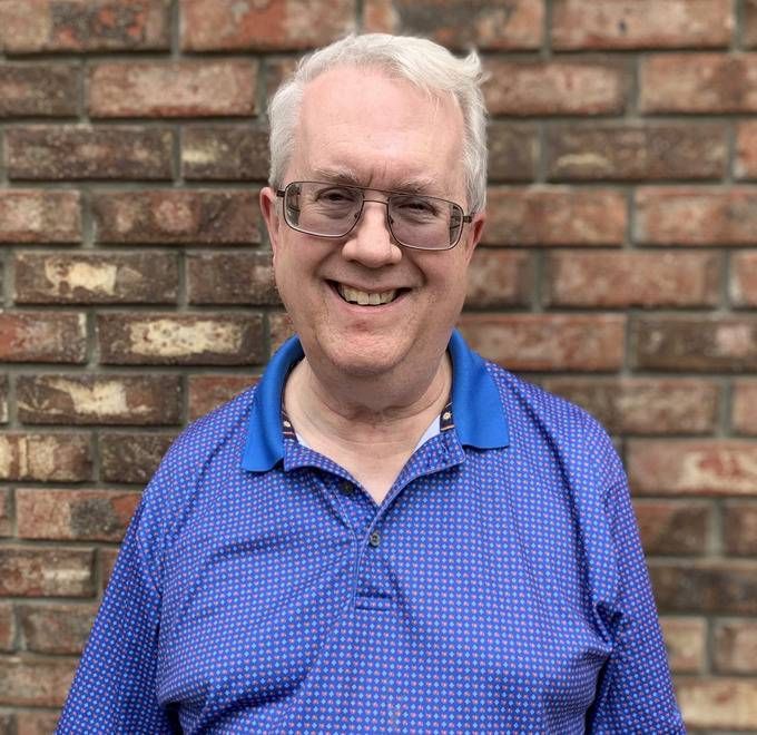 Smiling man in a blue polo shirt standing against a brick wall.