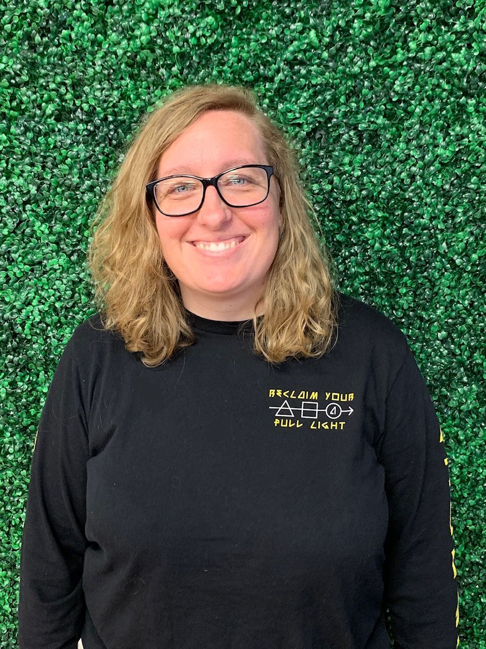 Smiling person with glasses and curly hair wearing a black shirt, standing before a green leafy backdrop