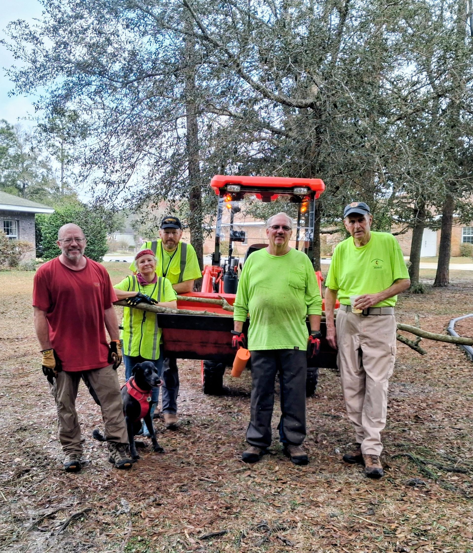 Five workers in bright safety shirts stand beside an orange utility vehicle in a wooded area.