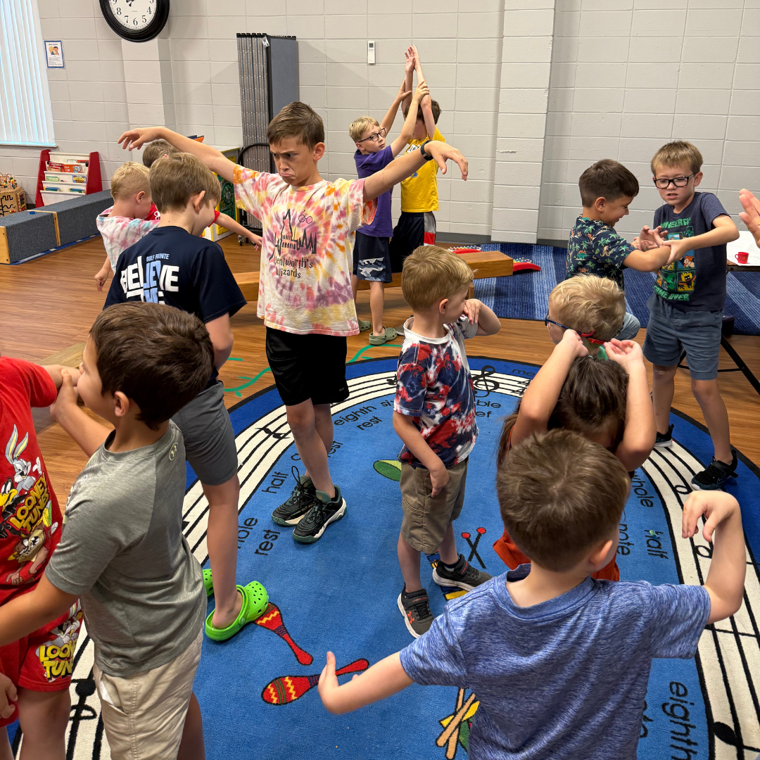 Children playing on a gym floor, some stretching with arms raised, in a school gymnasium.