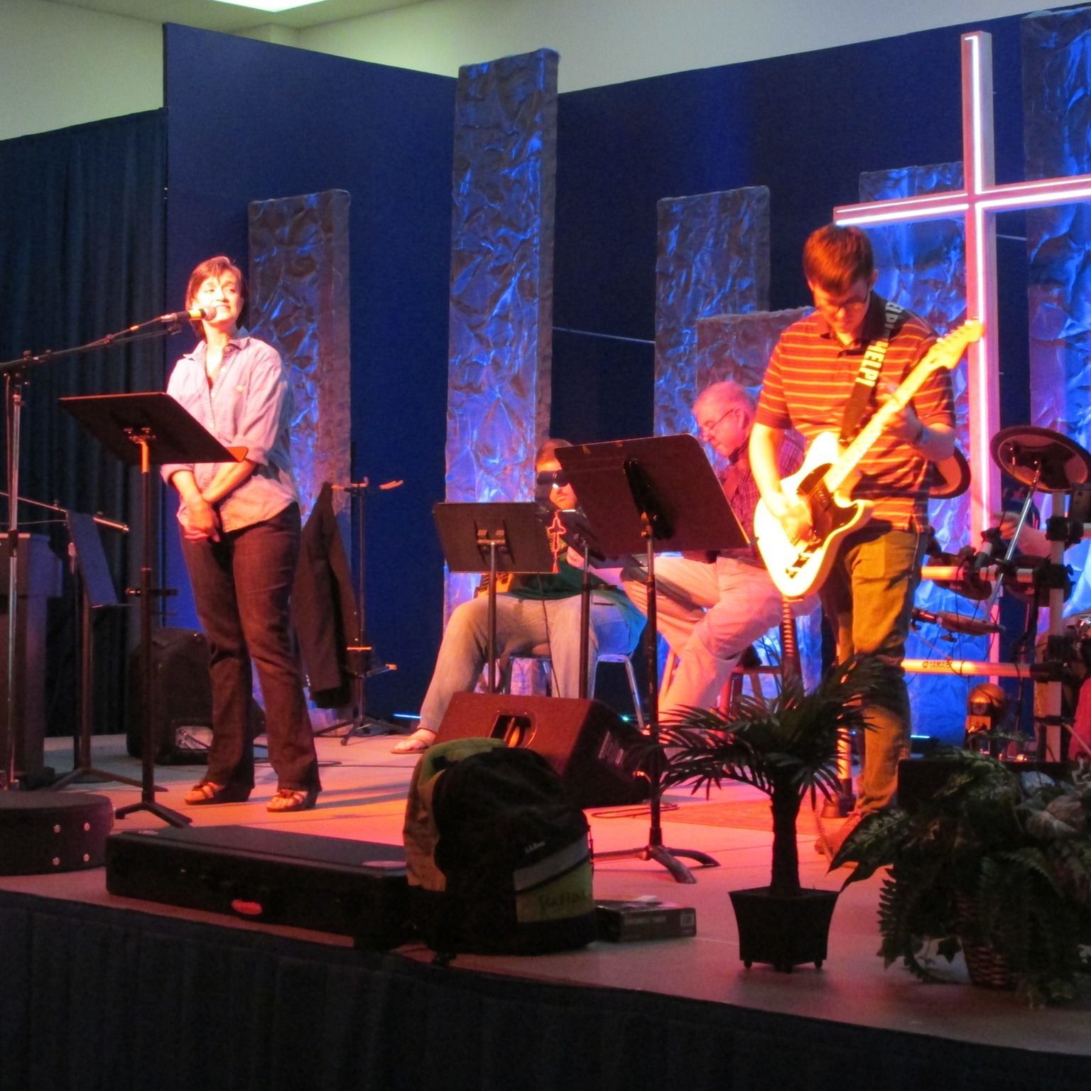 Musicians performing on a small stage with microphones, guitars, drums, and a glowing cross backdrop.