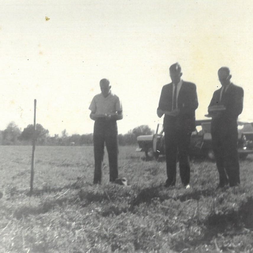 Three people stand in a grassy field beside a parked car, with a post marker in the foreground.