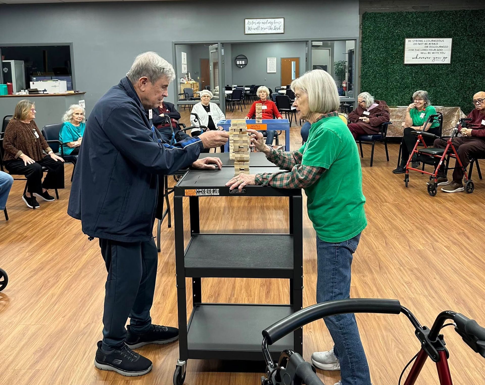 Two people play a giant tabletop game in a community room, with seated spectators around them.