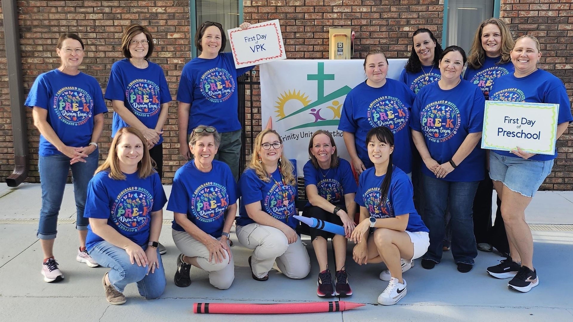 Group in blue shirts posing outdoors with signs and a cross mural behind them