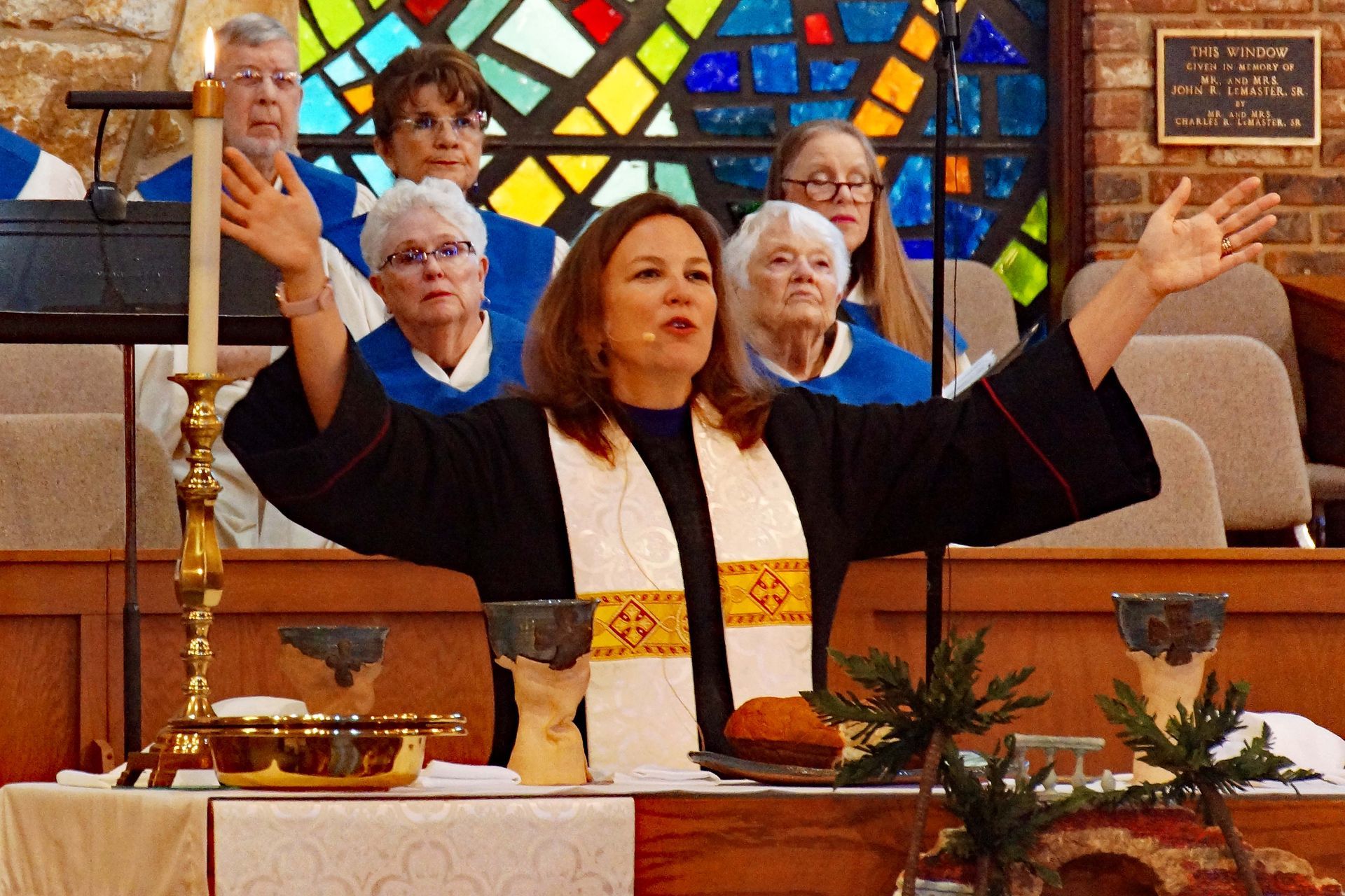 Woman leading worship at a church altar with arms raised, stained-glass windows behind her.