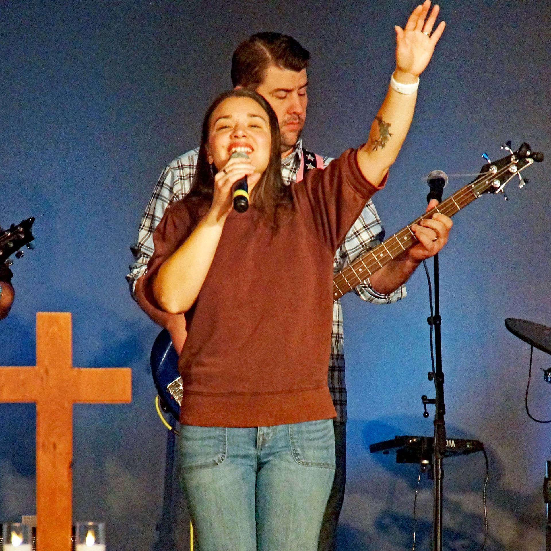 Singer holding a microphone with one arm raised onstage, with a guitarist behind her and a large cross at left.