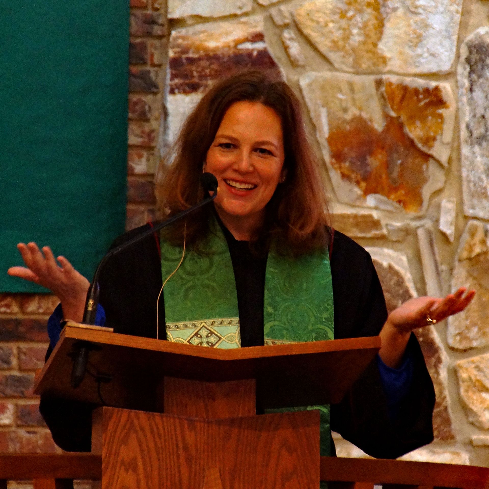 Woman speaking at a podium with a microphone, wearing a black robe and green stole, against a stone wall.