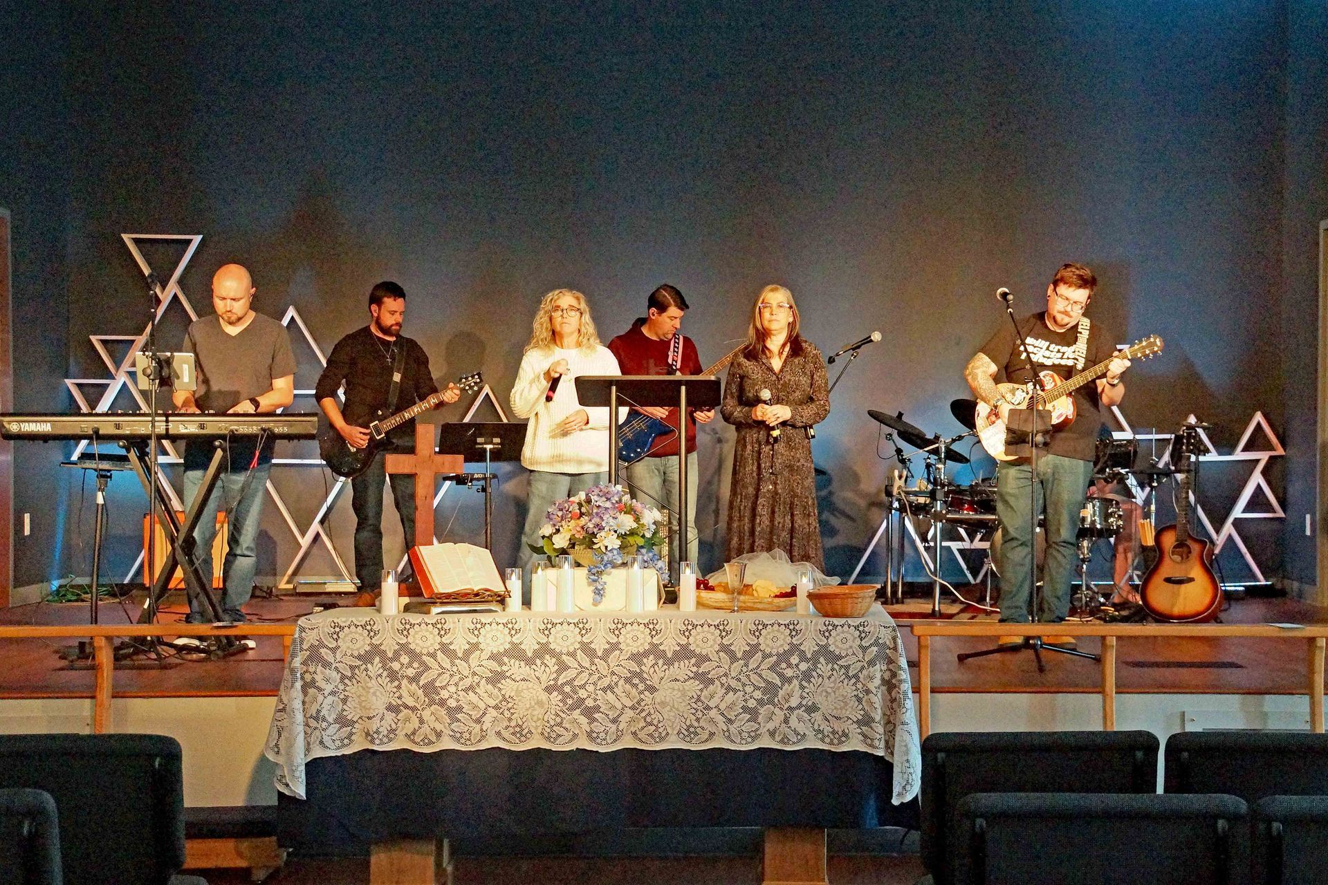 Band performing on a church stage with instruments and microphones behind a lace-covered table.