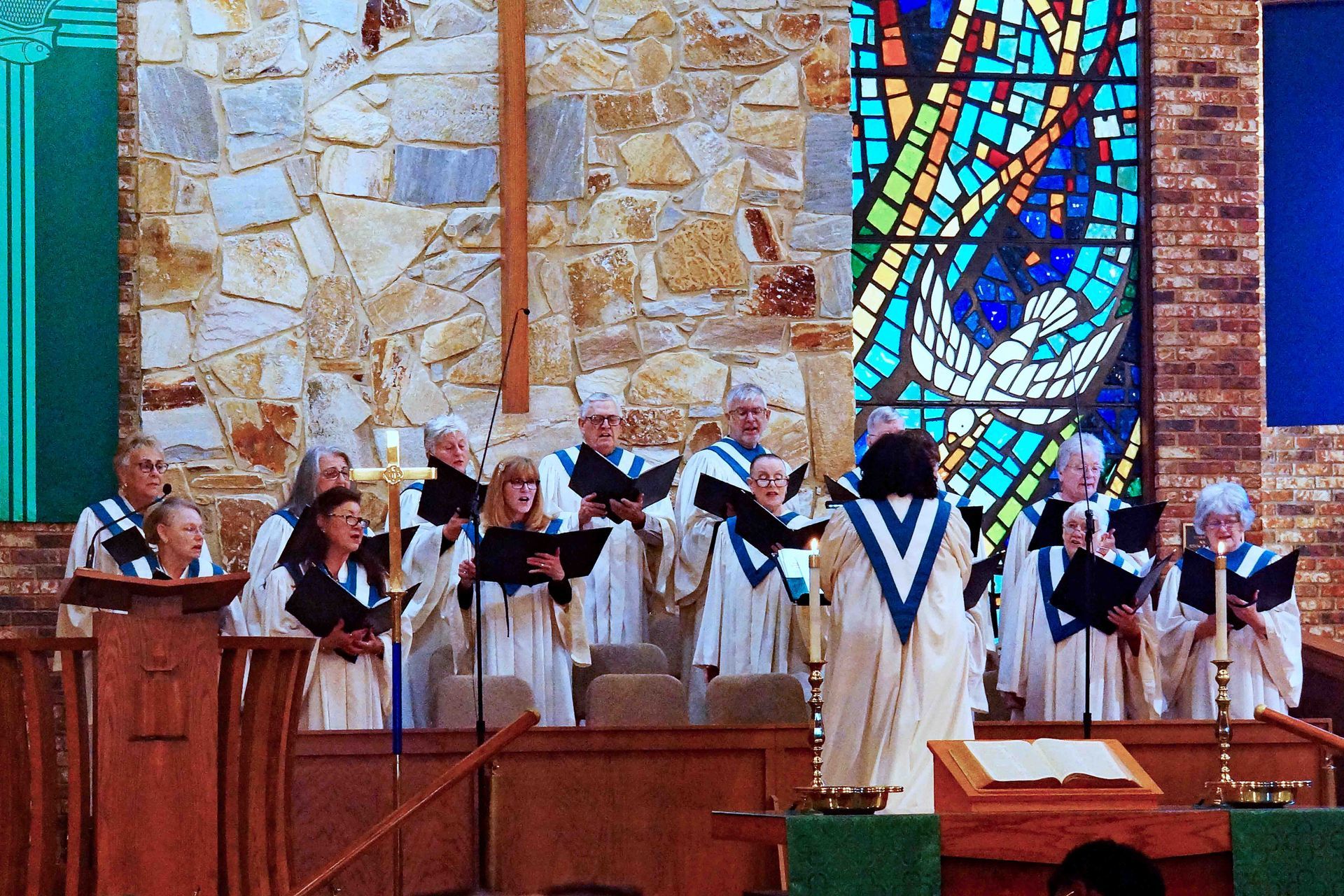 Choir in white robes singing in a church sanctuary before stained glass and stone walls