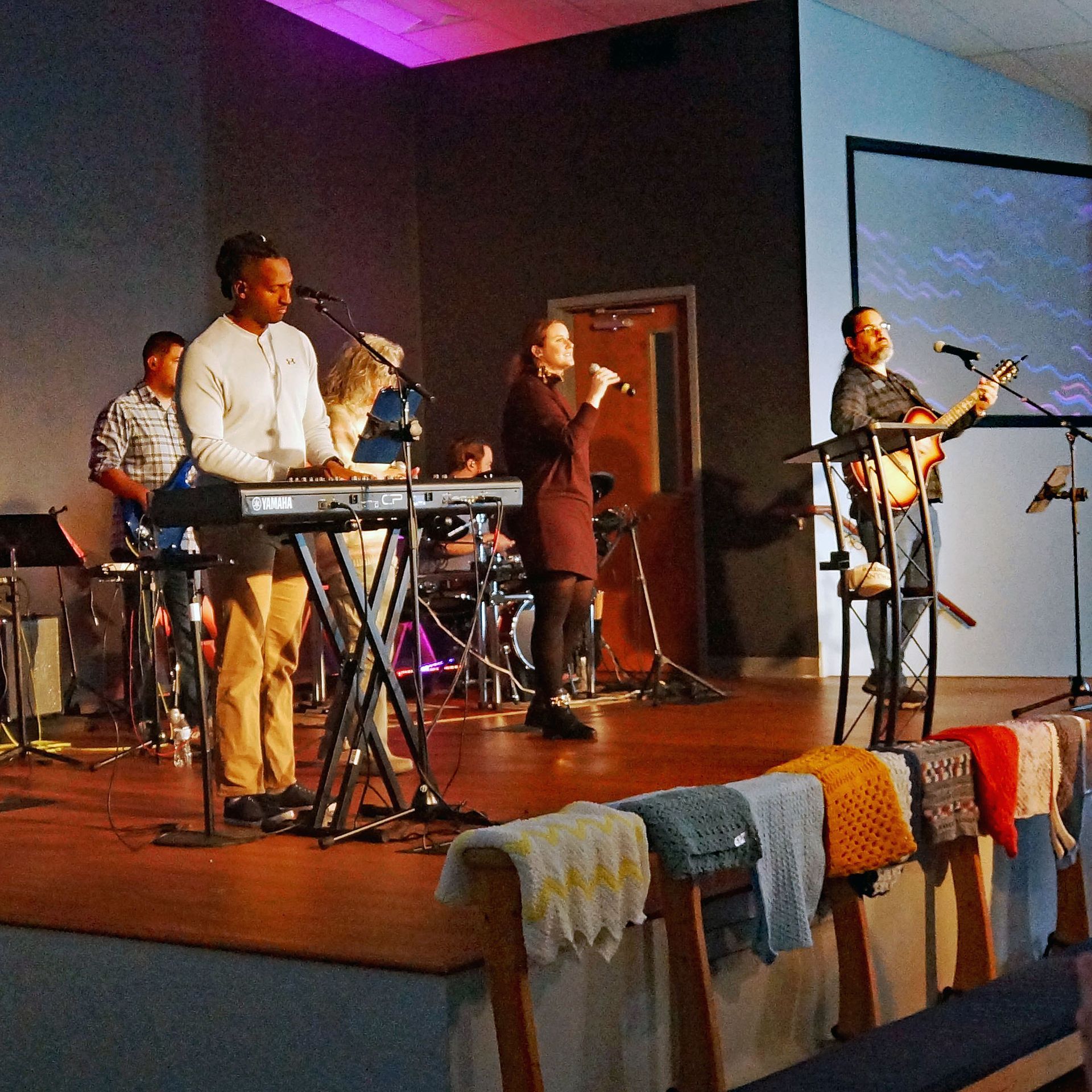Musicians performing on a church stage with keyboards, microphones, and colorful cloth draped over the front.