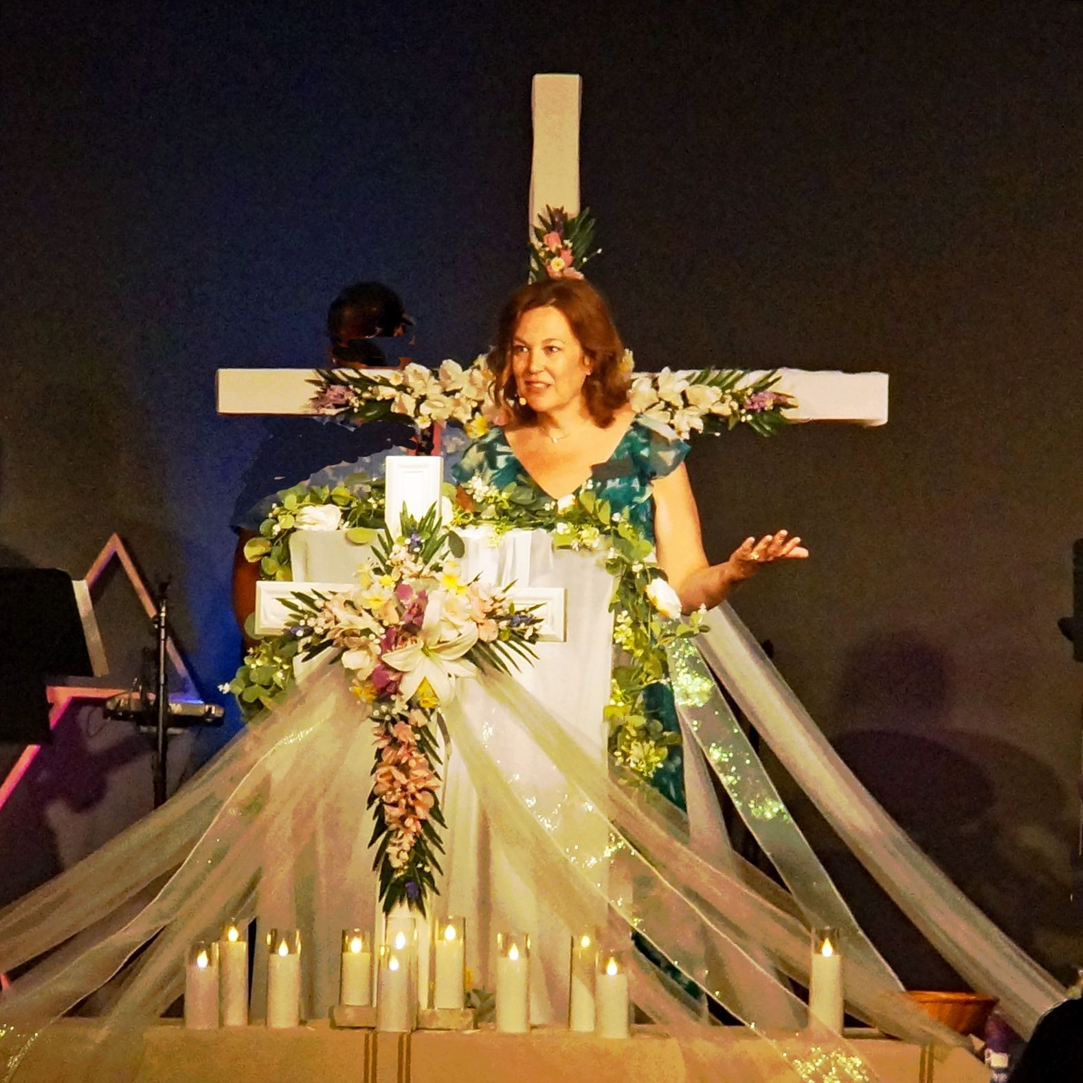 Woman at a decorated cross onstage, speaking with hands raised, surrounded by flowers and draped fabric.
