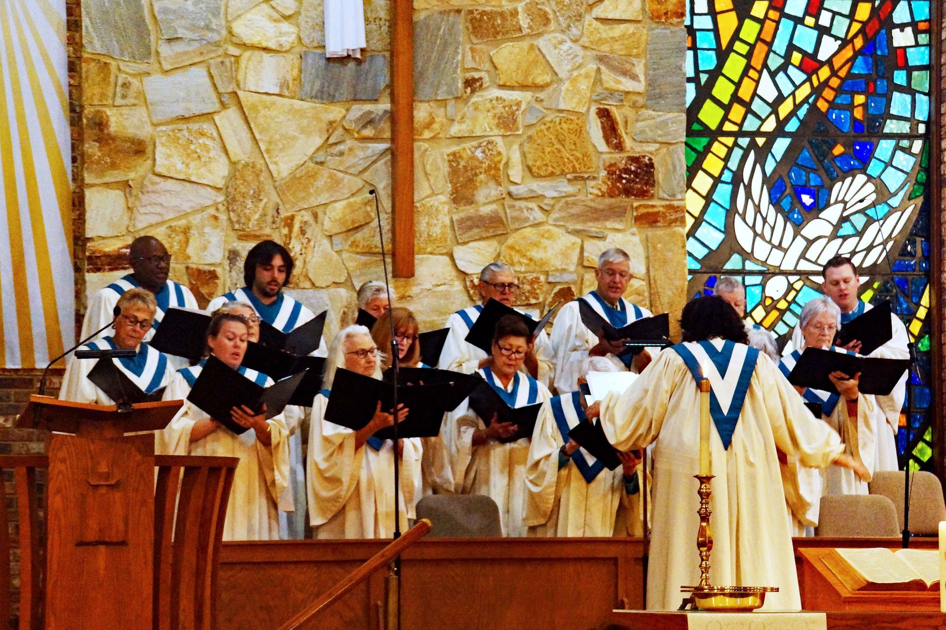 Choir in white robes sings with black folders in a church, standing before stone walls and stained glass.
