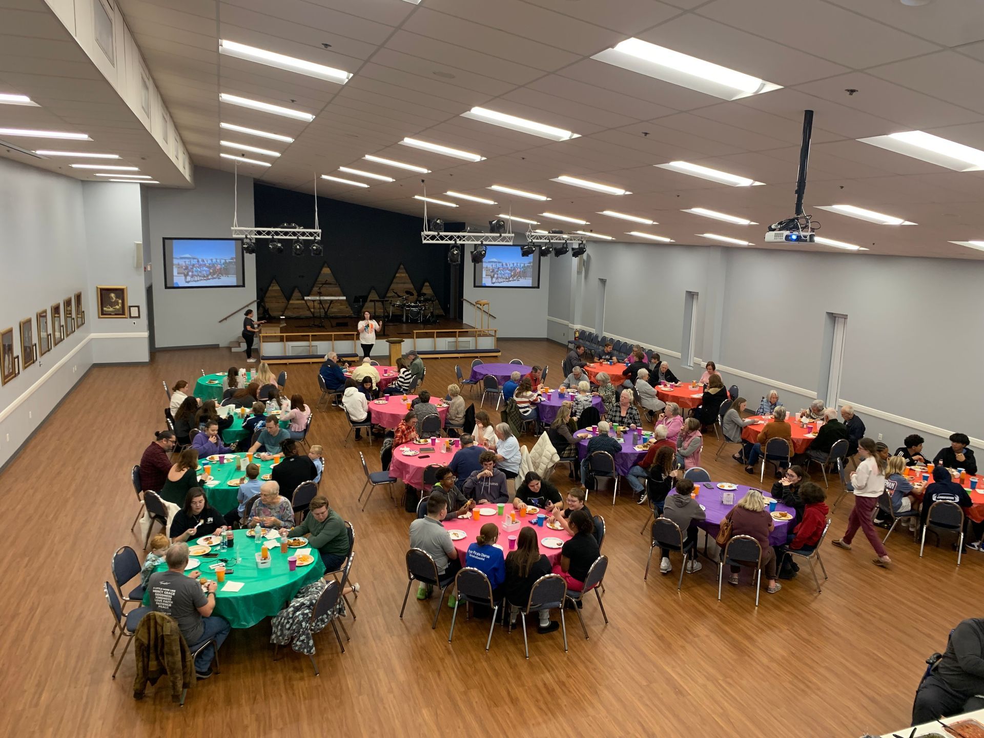 Large banquet hall with people seated at round tables, facing a stage with screens at the front.
