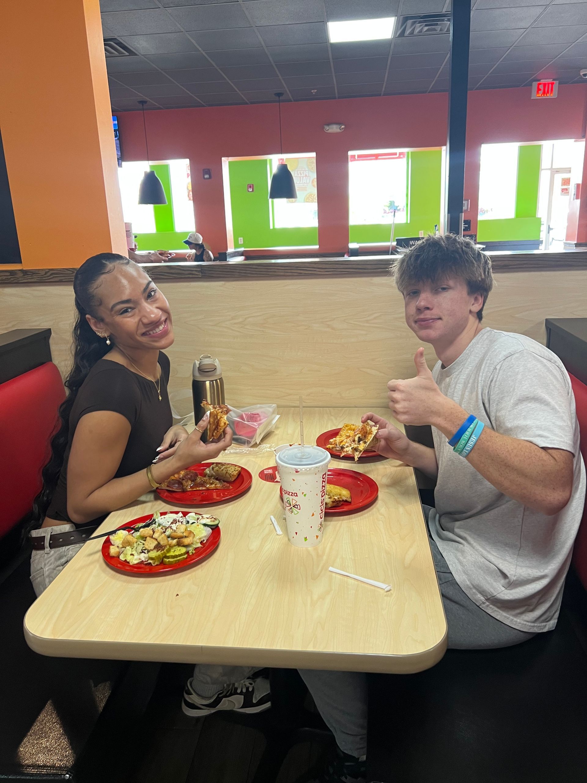 Two people sitting at a table in a restaurant, smiling and holding drinks with plates of food.
