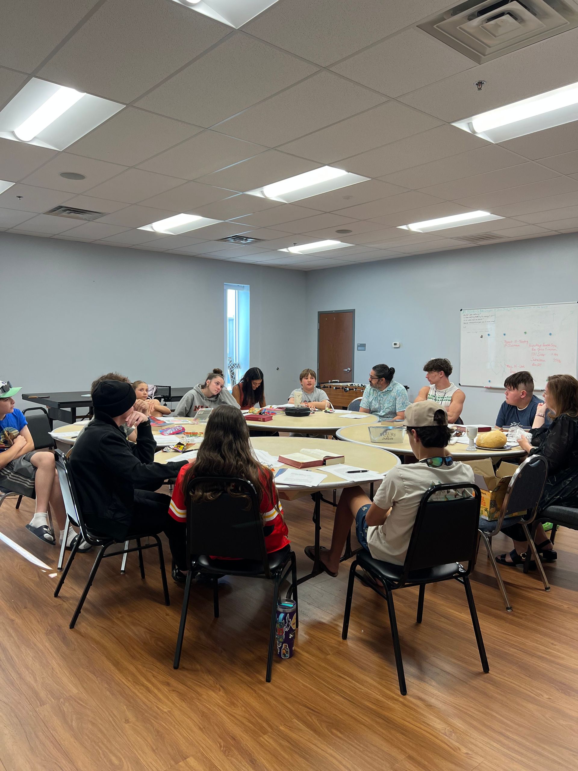 People seated around tables in a bright classroom meeting room, discussing papers and laptops