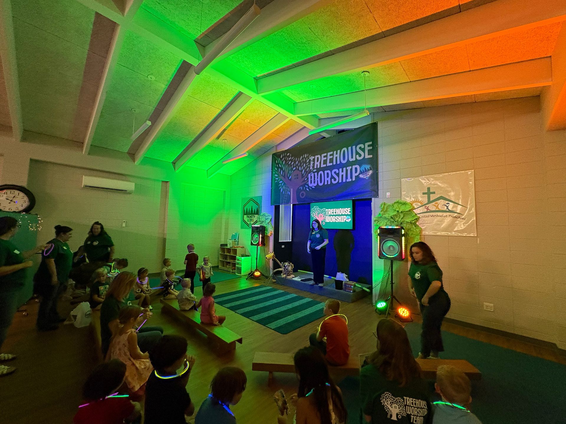 People gathered in a gym-like hall around a blue-lit stage with green and orange ceiling lights.