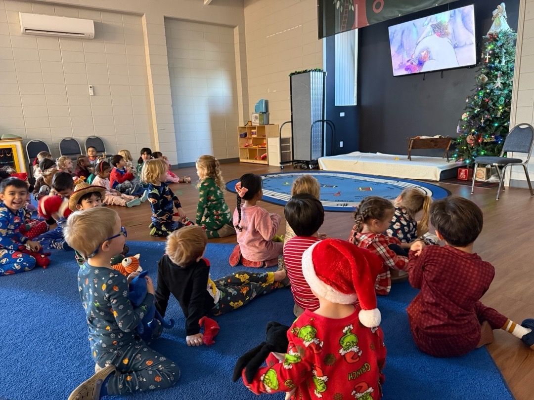 Children in pajamas sit on a classroom floor watching a holiday video on a screen.
